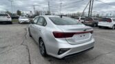 Rear view of a silver Kia Forte in a busy car dealership lot on a cloudy day while other vehicles surround it.