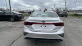 Rear view of a white Kia Forte in a car dealership lot, with several other cars and an overcast sky above.