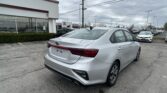 White Kia sedan in a dealership lot with other cars and a building in the background under a cloudy sky.