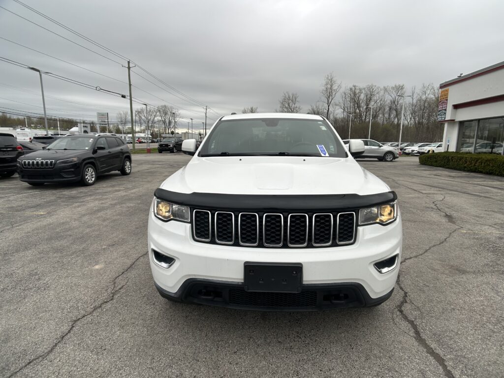White SUV seen from the front in a dealership lot with other cars and a gray overcast sky behind it.