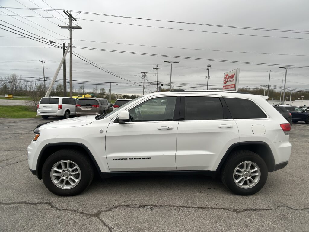 White Jeep Grand Cherokee in a parking lot with overhead power lines and a red EZ LOAN Auto Sales sign in the background, daytime.