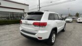 White Jeep SUV (Laredo) in a dealership lot with a white building to the left and rows of cars under a gray, cloudy sky.