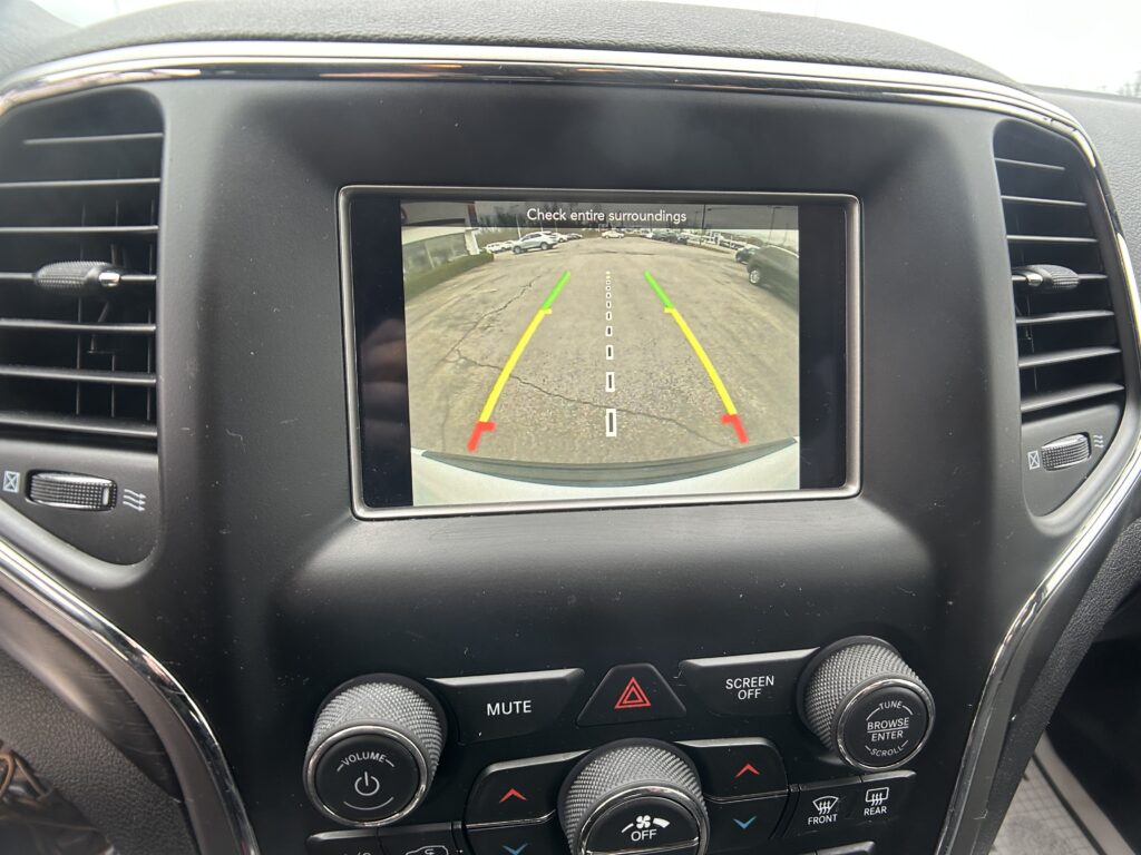Car infotainment screen showing rearview camera view with parking lot, guiding lines, and the message 'Check entire surroundings'