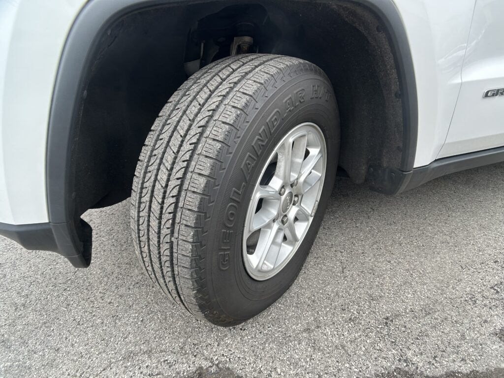 Front wheel of a light gray SUV, showing a treaded tire in the wheel well with a silver alloy rim