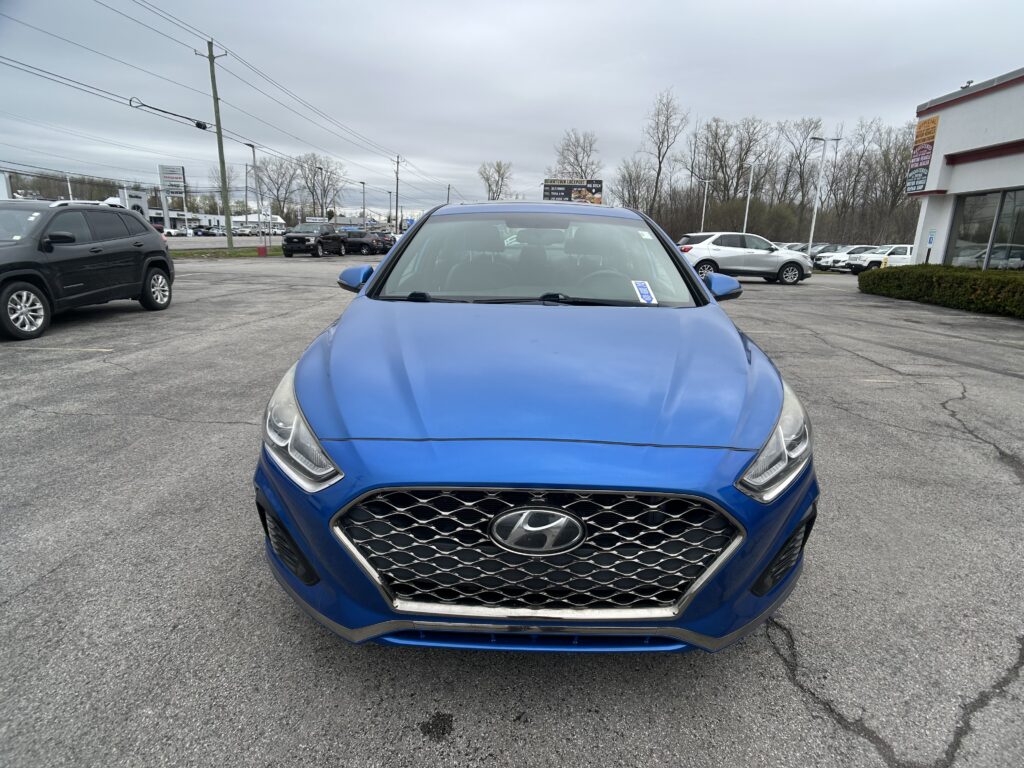 Front view of a blue Hyundai SUV in a car dealership lot, with other cars and a building in the background