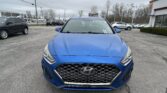 Front view of a blue Hyundai SUV in a car dealership lot, with other cars and a building in the background