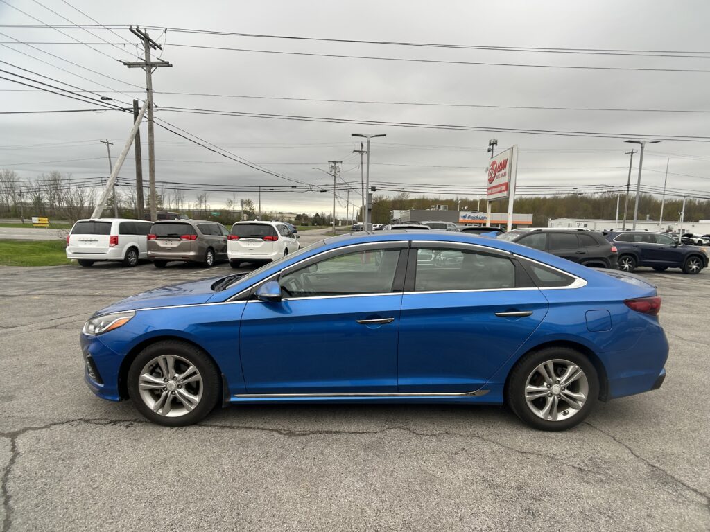 Blue sedan in the foreground of a busy parking lot with white vans and other cars, overcast sky and many power lines overhead.