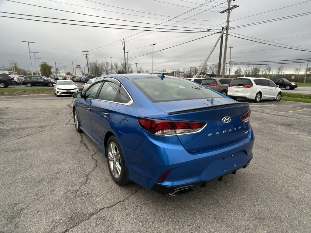 Blue Hyundai Sonata parked in a dealership lot, shown from the rear-right with other cars and overhead power lines under a cloudy sky,