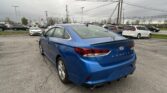 Blue Hyundai Sonata parked in a dealership lot, shown from the rear-right with other cars and overhead power lines under a cloudy sky,