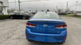 Rear view of a blue Hyundai Sonata Sport in a car dealership lot, with other vehicles and a white building nearby.