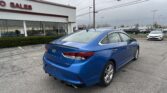 Blue Hyundai Sonata parked in a dealership lot with a white building labeled SALES on the left under an overcast sky.