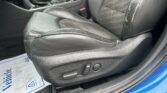 Close-up of a black leather car driver’s seat with lumbar/seat adjustment controls and red seatbelt buckle, blue door sill visible outside.