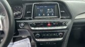 View of a car interior from the driver’s seat, showing the steering wheel, instrument cluster, and a center touchscreen displaying a radio (91.7) with car controls and icons, plus climate controls below. Outside parking lot visible through windshield.