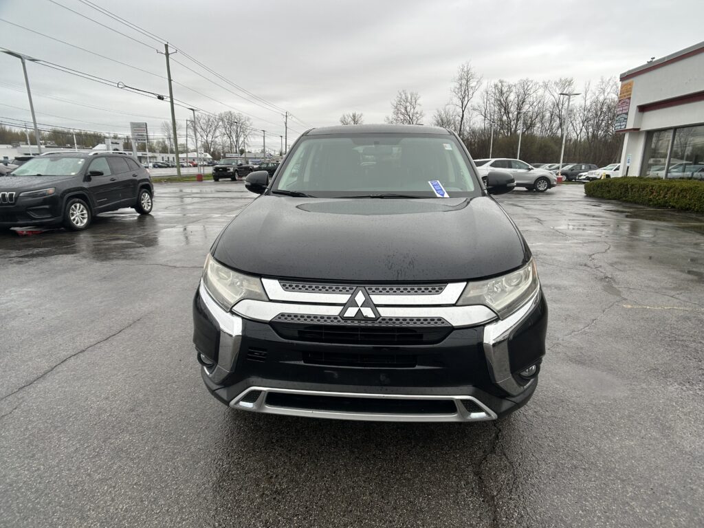 Front view of a dark Mitsubishi SUV in a rainy dealership lot, with a showroom building to the right and other cars in the background.