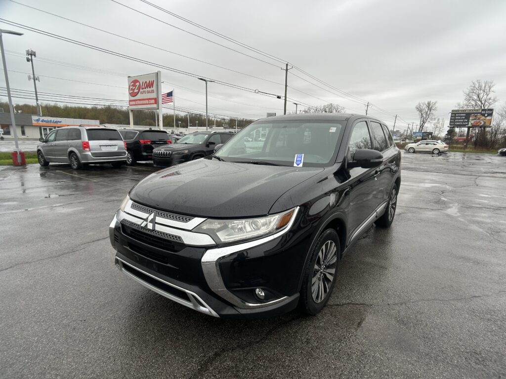 Close-up of a black Mitsubishi SUV in a wet parking lot with other vehicles and an EZ Loan sign in the background on an overcast day.