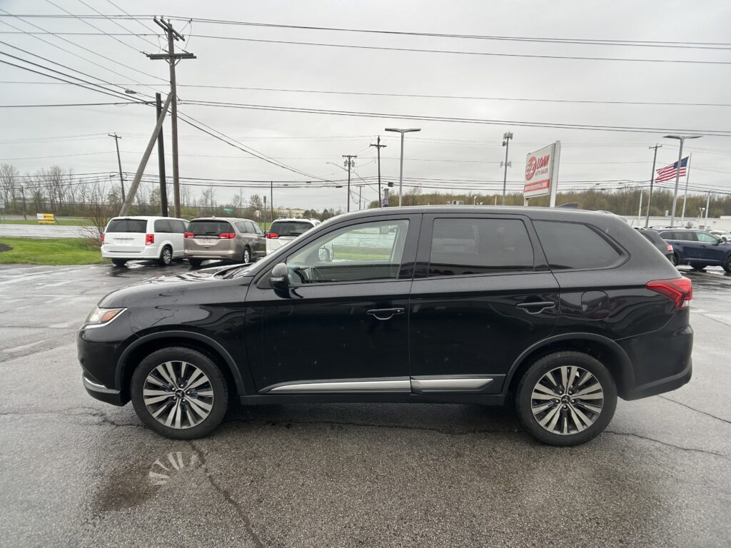 Black SUV in foreground of a car dealership lot, with white minivans behind and an American flag in the distance on an overcast day.
