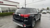 Black Mitsubishi Outlander SE SUV in a dealership lot, rain droplets on the vehicle, storefront in background.