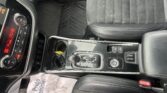 Front interior of a car showing the center console with gear shifter, cup holder, and black leather seats with white stitching on the right side of the image.