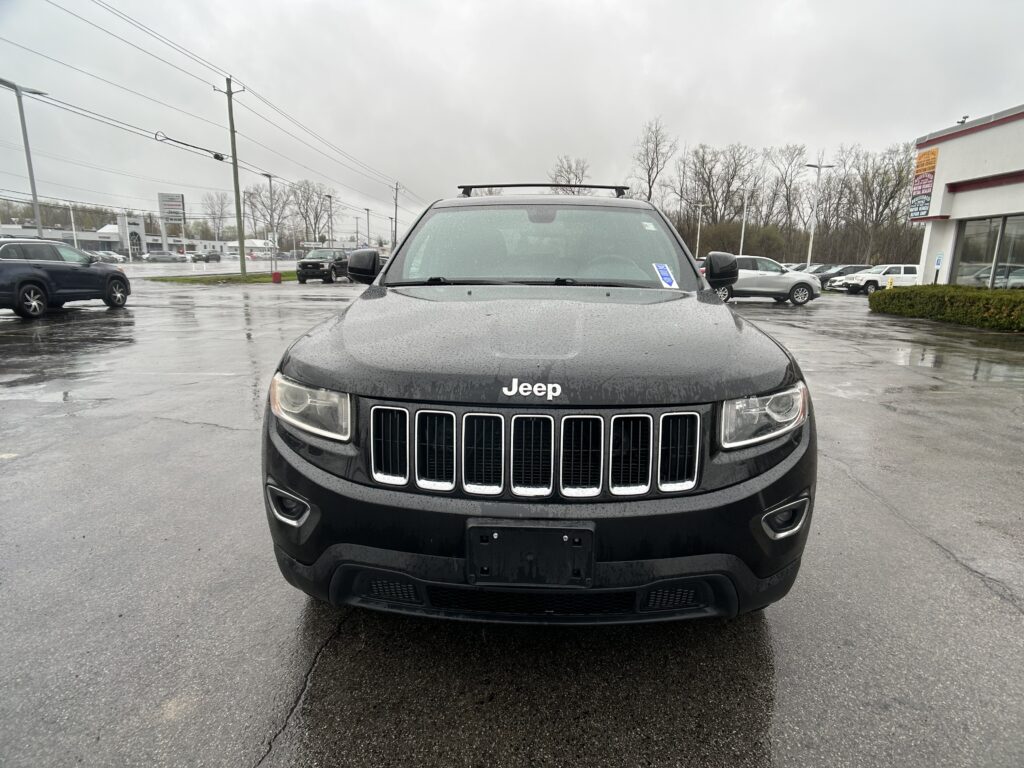 Front view of a black Jeep SUV with rain droplets on the hood, parked in a wet dealership lot with buildings in the background.