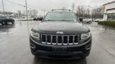 Front view of a black Jeep SUV with rain droplets on the hood, parked in a wet dealership lot with buildings in the background.