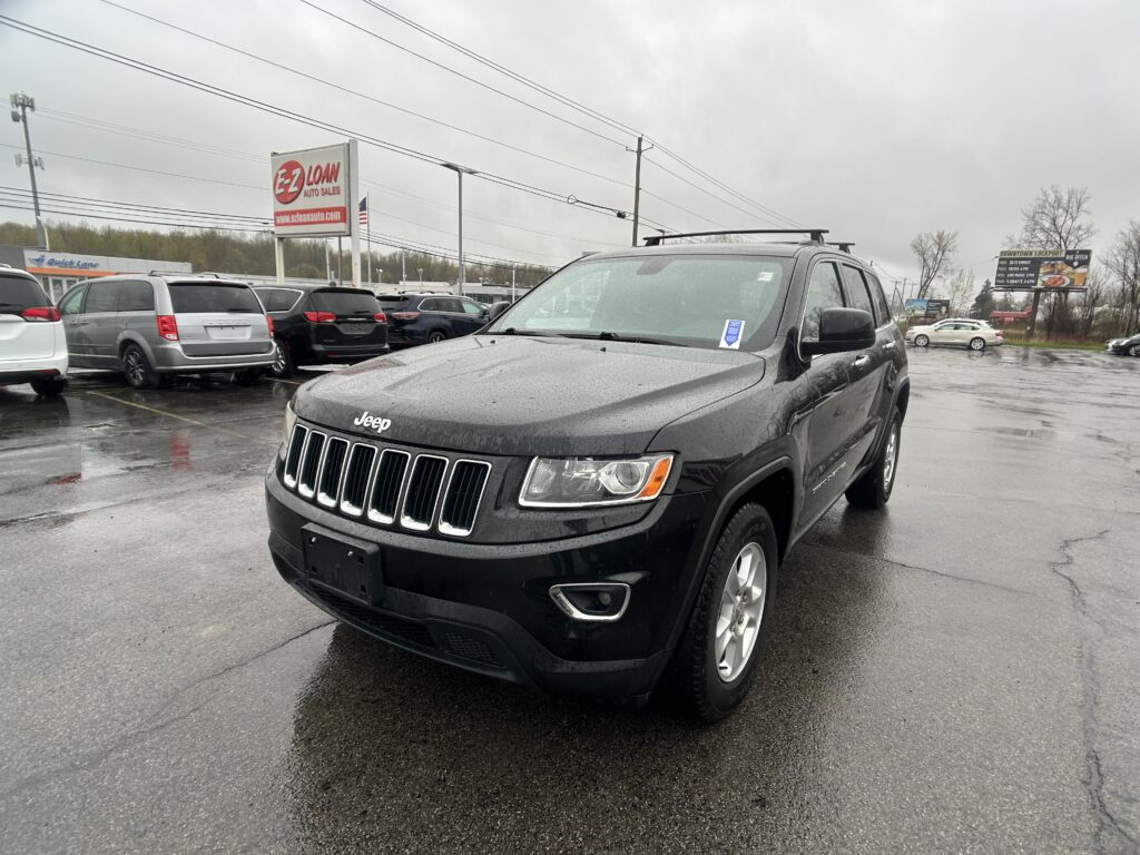 Gray Jeep SUV parked in a rainy parking lot with wet pavement and other cars in the background, advertising signs for auto sales in the distance.