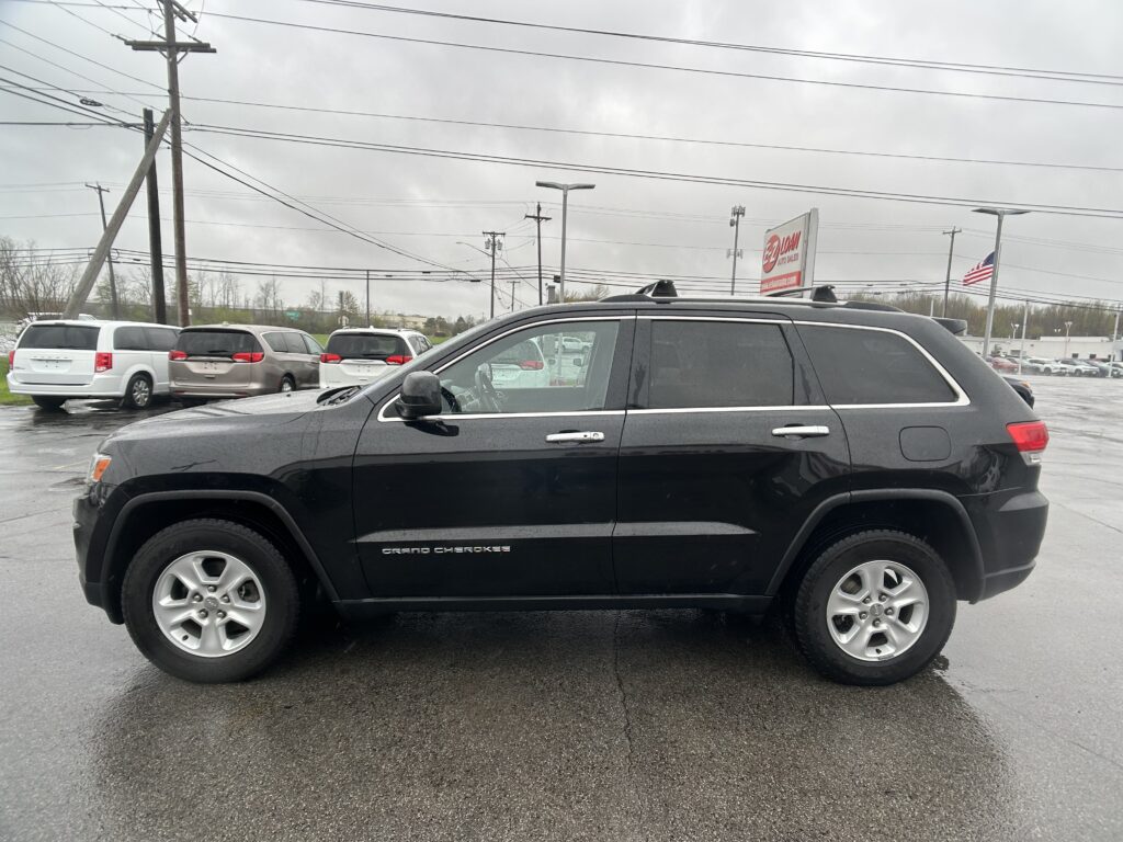 Black Grand Cherokee SUV in foreground at a car dealership lot on a wet day, with a service sign and power lines in the background
