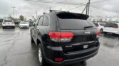 Black Jeep SUV (4x4) photographed from the rear in a rainy dealership lot with other cars and power lines overhead.