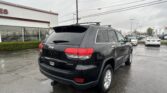 Back-right three-quarter view of a black Jeep Grand Cherokee with roof rails in a rainy dealership lot, with other cars and a white building nearby.