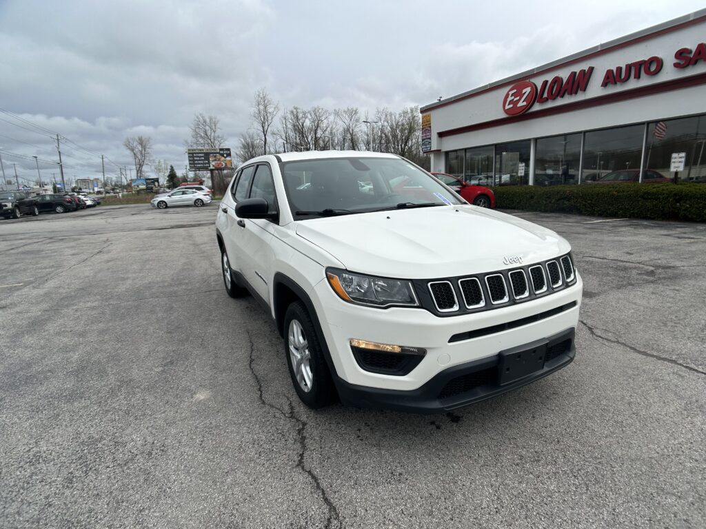 White Jeep SUV parked in a dealership lot in front of a glass-walled showroom with an EZ Loan Auto sign on the building, under a cloudy sky.