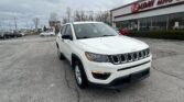 White Jeep SUV parked in a dealership lot in front of a glass-walled showroom with an EZ Loan Auto sign on the building, under a cloudy sky.