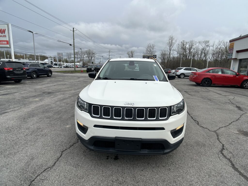 Front view of a white Jeep SUV in a car dealership lot, with other cars and a cloudy sky in the background, grille and headlights visible.