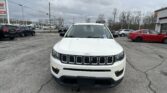 Front view of a white Jeep SUV in a car dealership lot, with other cars and a cloudy sky in the background, grille and headlights visible.