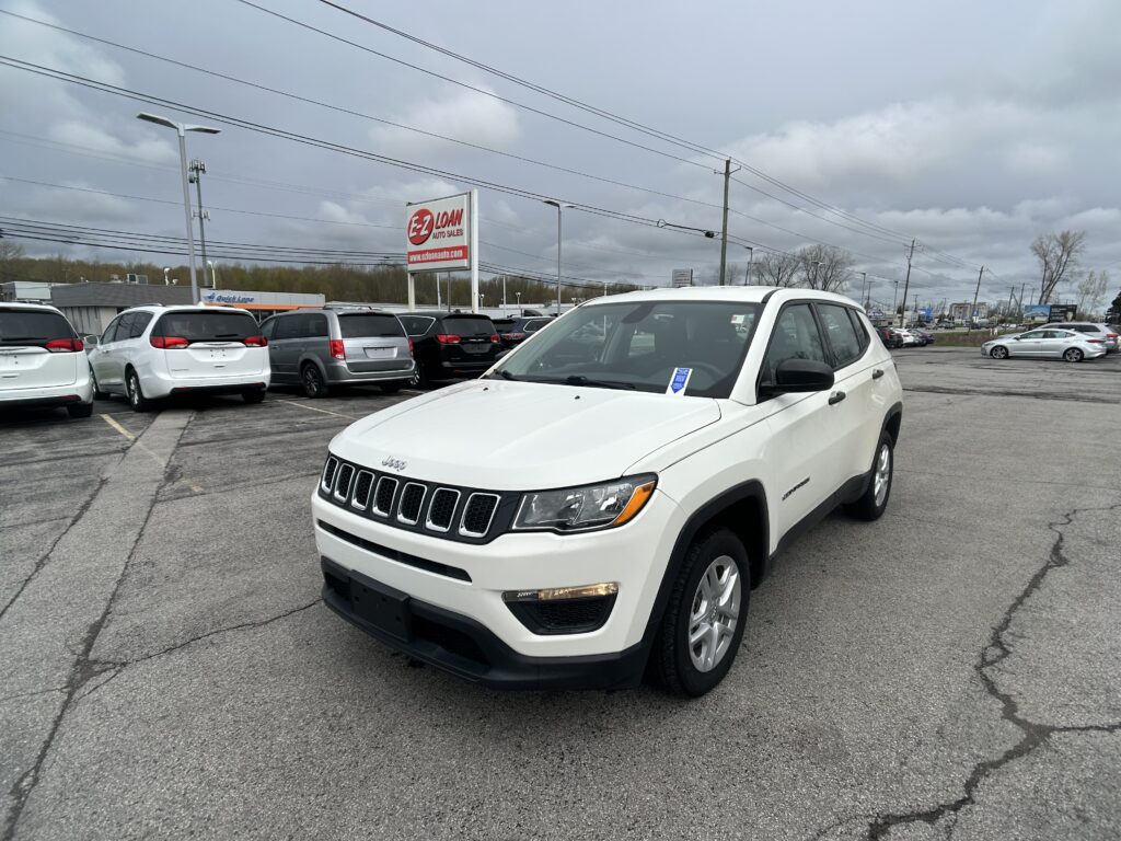 White Jeep SUV in a dealership lot with other minivans and an EZ Loan sign under a cloudy sky.