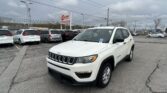 White Jeep SUV in a dealership lot with other minivans and an EZ Loan sign under a cloudy sky.