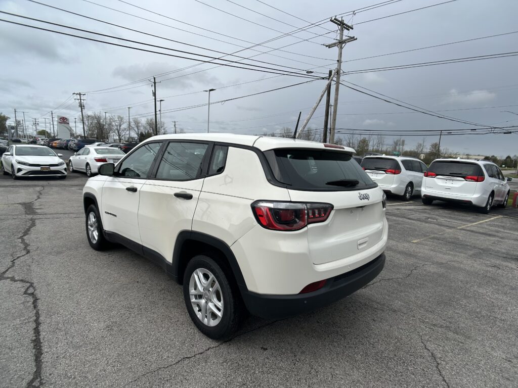 White Jeep SUV in the foreground of a car dealership lot, with multiple white cars behind it and power lines overhead under a cloudy sky.