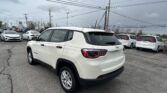White Jeep SUV in the foreground of a car dealership lot, with multiple white cars behind it and power lines overhead under a cloudy sky.