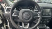 Steering wheel and dashboard of a Jeep, with instrument cluster and center console visible; parking lot outside through windshield.