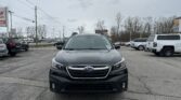 Front view of a dark gray Subaru SUV in a parking lot, with other cars and bare trees under a cloudy sky.