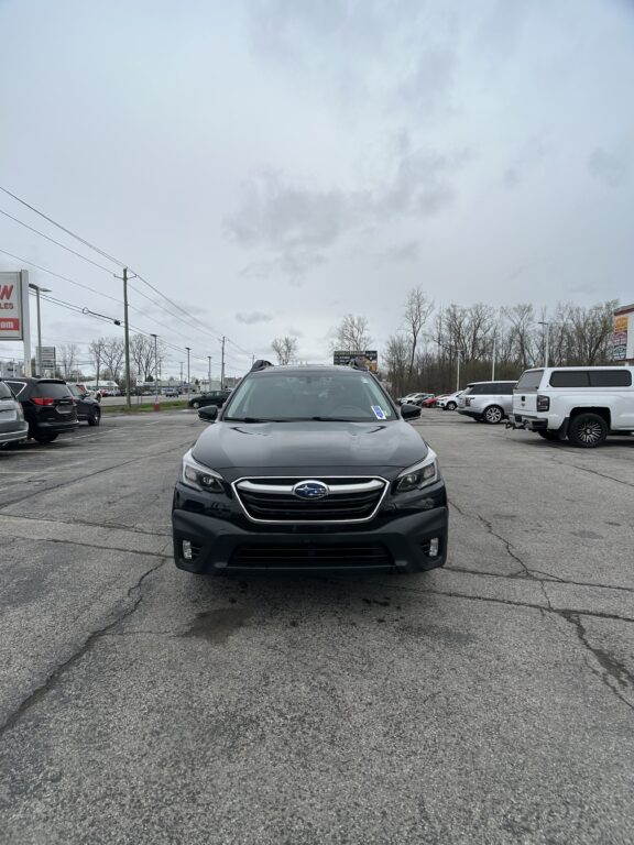 Front view of a dark gray Subaru SUV in a parking lot, with other cars and bare trees under a cloudy sky.