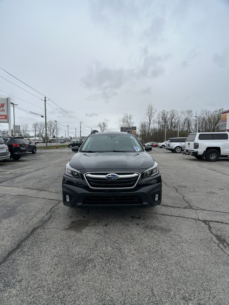Front view of a dark gray Subaru SUV in a parking lot, with other cars and bare trees under a cloudy sky.