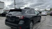 Black Subaru Outback SUV shown from the rear in a car dealership lot, with other cars and power lines overhead under a cloudy sky.
