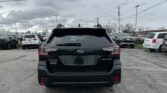 Rear view of a black Subaru Outback in a parking lot, with other cars and power lines overhead under a cloudy sky.