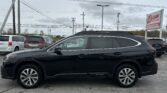 Black Subaru Outback SUV in a dealership lot with other cars and an EZ LOAN Auto Sales sign in the background, under cloudy skies.