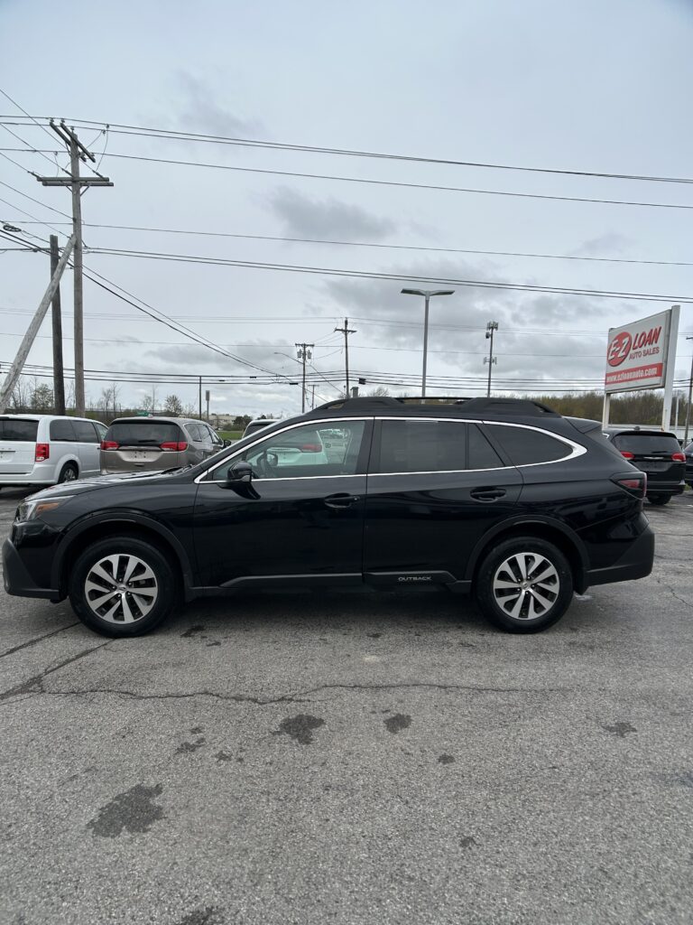 Black Subaru Outback SUV in a dealership lot with other cars and an EZ LOAN Auto Sales sign in the background, under cloudy skies.