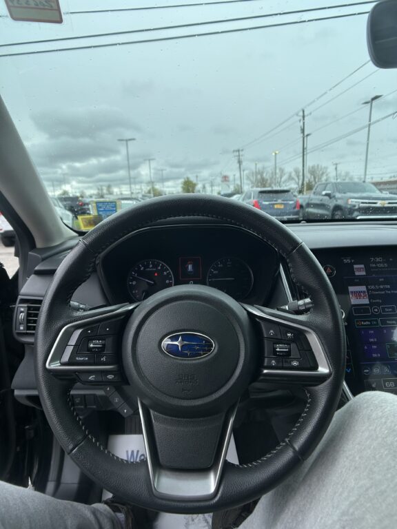 Driver's view from the driver's seat showing the steering wheel, instrument cluster, and a rain-speckled windshield with a parking lot outside.