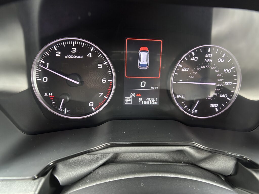 Center dashboard display showing a top-down car icon inside a red outline (parking status), flanked by a tachometer on the left and a speedometer on the right; the odometer reads 115,610 miles.