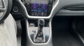 Interior of a car showing a large touchscreen infotainment system in the center console, gear shifter, and cup holders nearby, with a parking lot visible through the windshield.