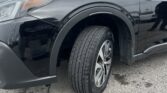 Close-up of the front left side of a dark SUV, showing the headlight, wheel, and fender with raindrops on the body. A cloudy sky and parking lot are in the background.