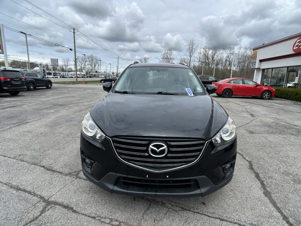Front view of a black Mazda SUV in a car dealership lot under a cloudy sky, with other cars and a building in the background.