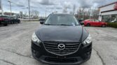 Front view of a black Mazda SUV in a car dealership lot under a cloudy sky, with other cars and a building in the background.